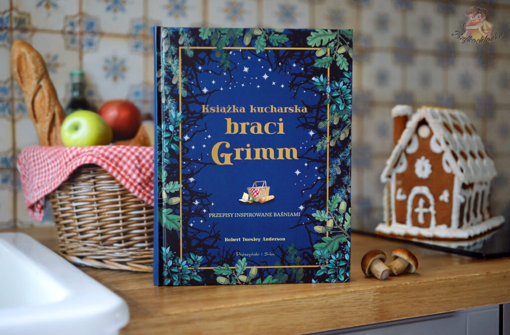 Okładka książki „Książka kucharska braci Grimm” z koszykiem pełnym jedzenia i domkiem z piernika inspirowanym baśnią Jasia i Małgosi.