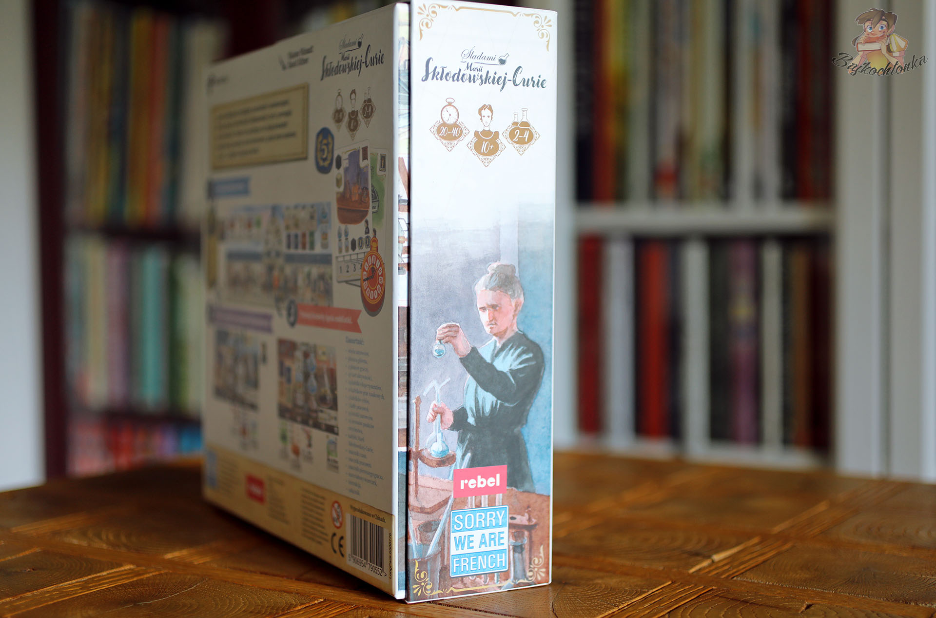 Bok pudełka gry Śladami Marii Skłodowskiej-Curie z ilustracją Marii podczas eksperymentu