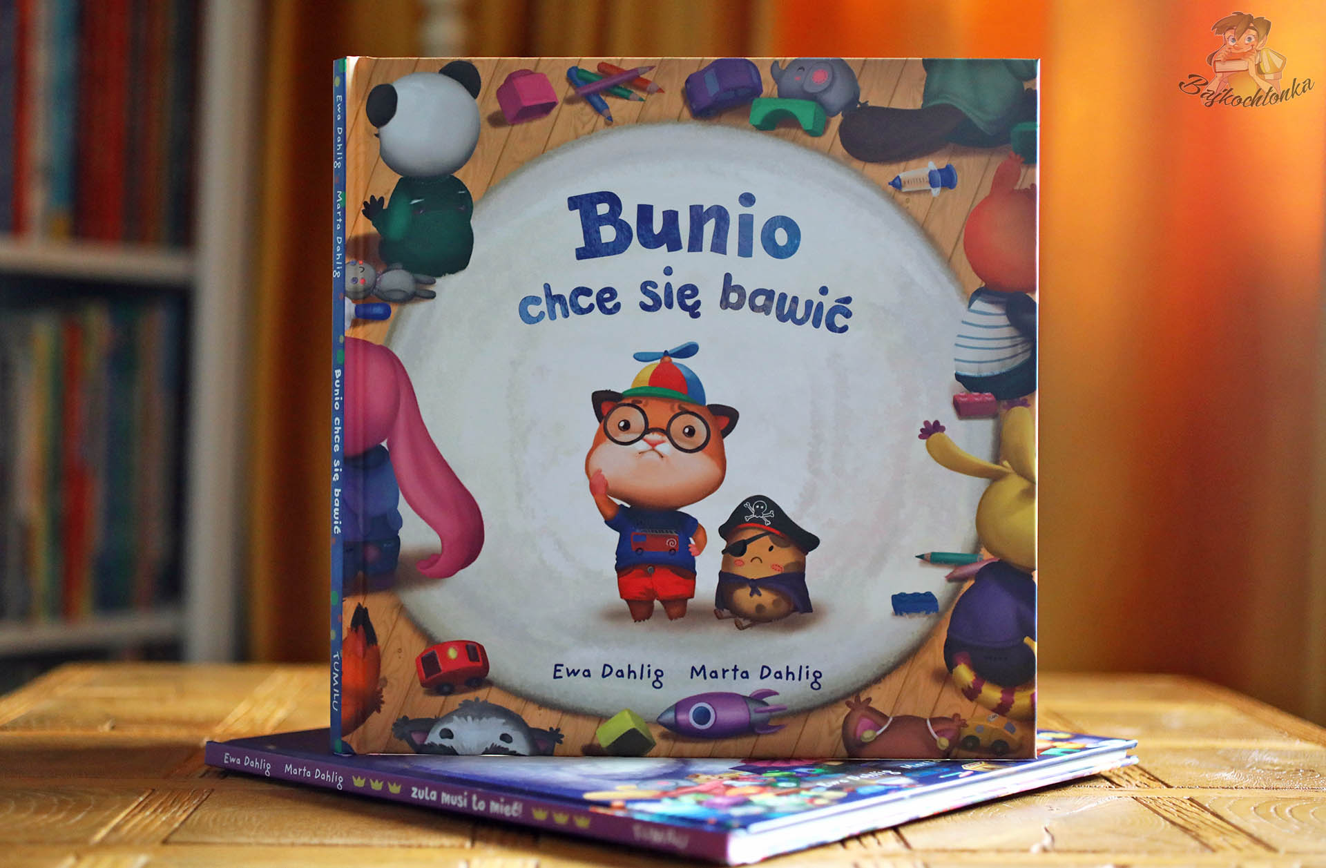 Książka „Bunio chce się bawić” Ewy i Marty Dahlig na tle półek – sympatyczny bohater w czapce z wirnikiem, lektura o przyjaźni w przedszkolu.