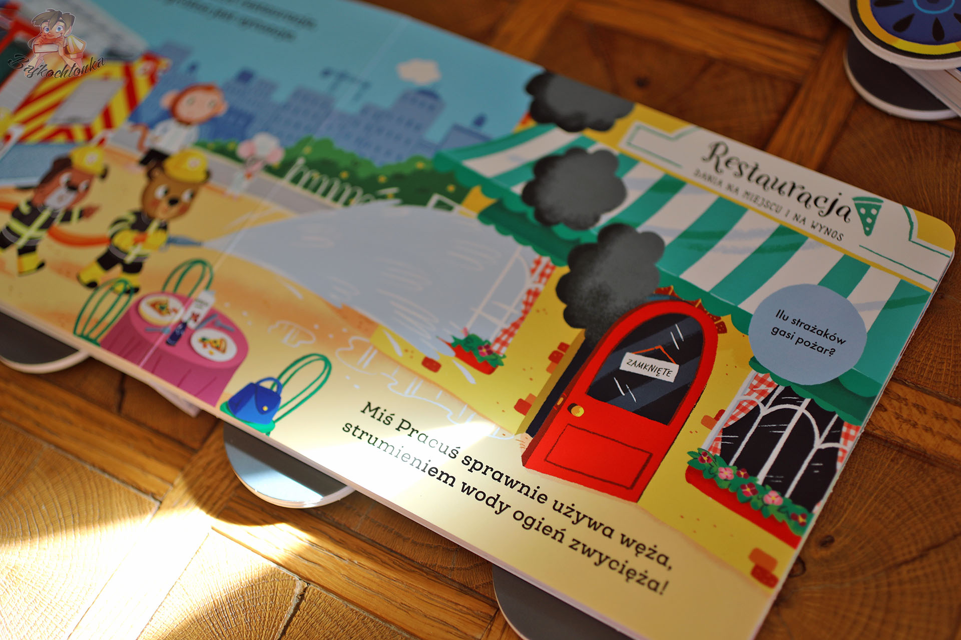 Rozkładówka z książki „Miś Pracuś. Wóz strażacki” – gaszenie pożaru przy restauracji, rymowany tekst, książka dla maluchów.