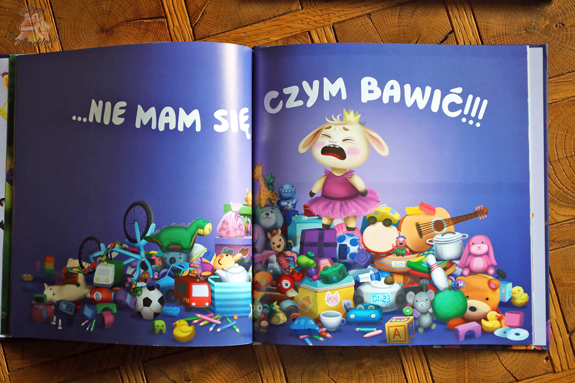 Dwustronicowa rozkładówka z „Zula musi to mieć!” – Zula krzyczy wśród góry zabawek, przesłanie o nadmiarze i mądrym kupowaniu.