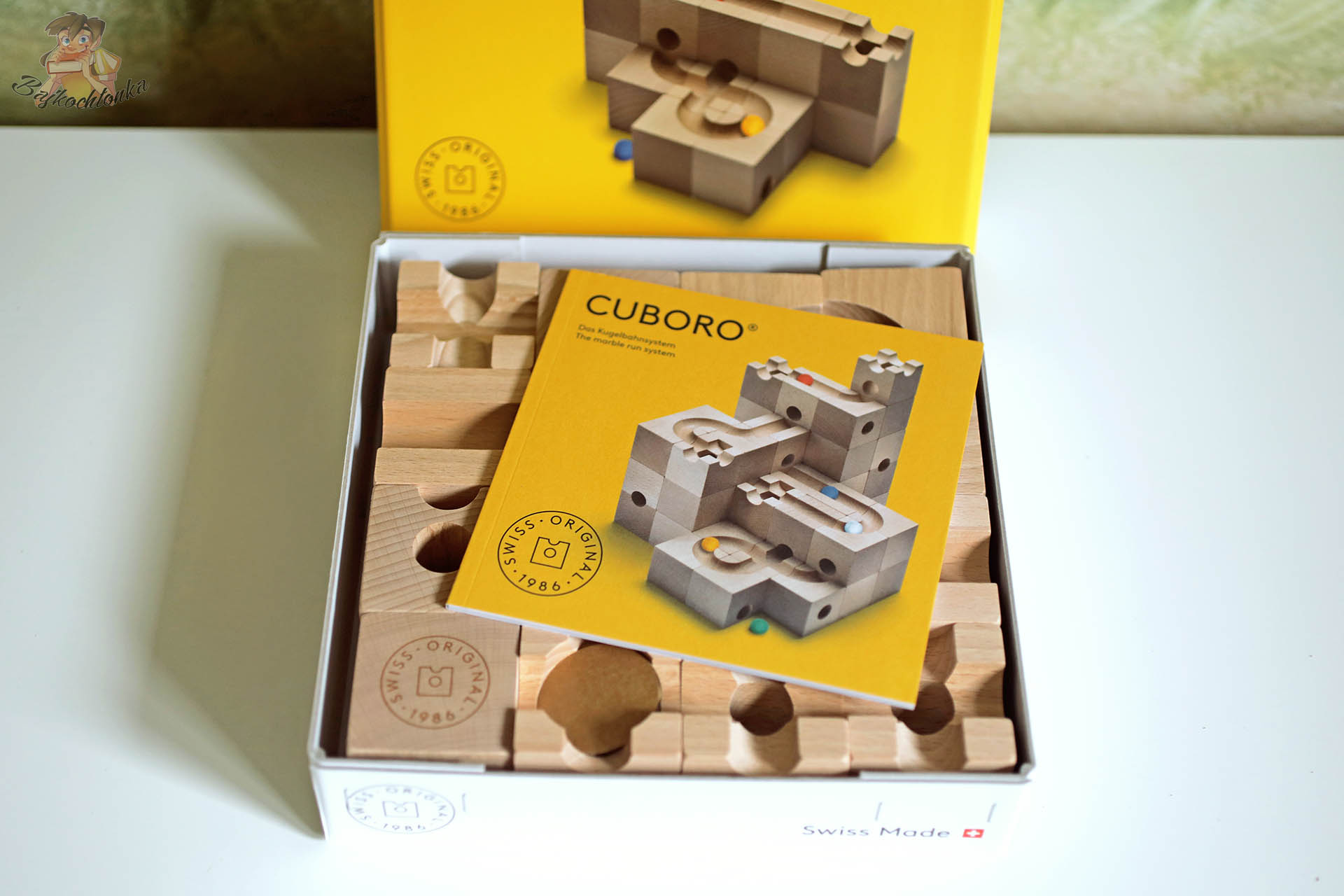 Otwarty karton z klockami Cuboro i żółtą instrukcją z ilustracją drewnianego toru kulkowego