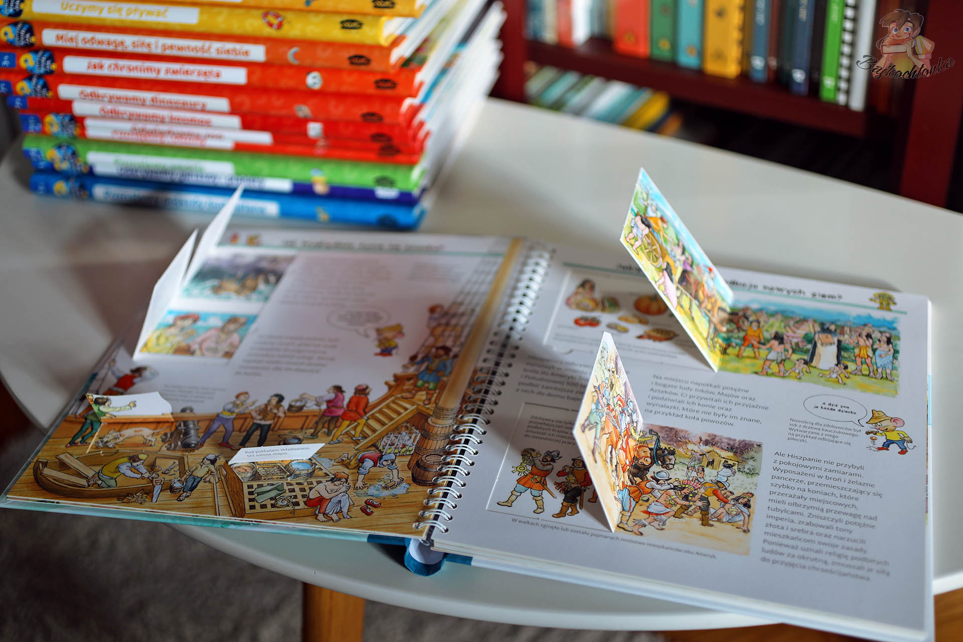 Otwarta książka Odkrywcy i podróżnicy z serii Dlaczego? Kiedy? Jak? – kolorowa rozkładówka historyczna z licznymi okienkami oraz stos innych książek z serii w tle.