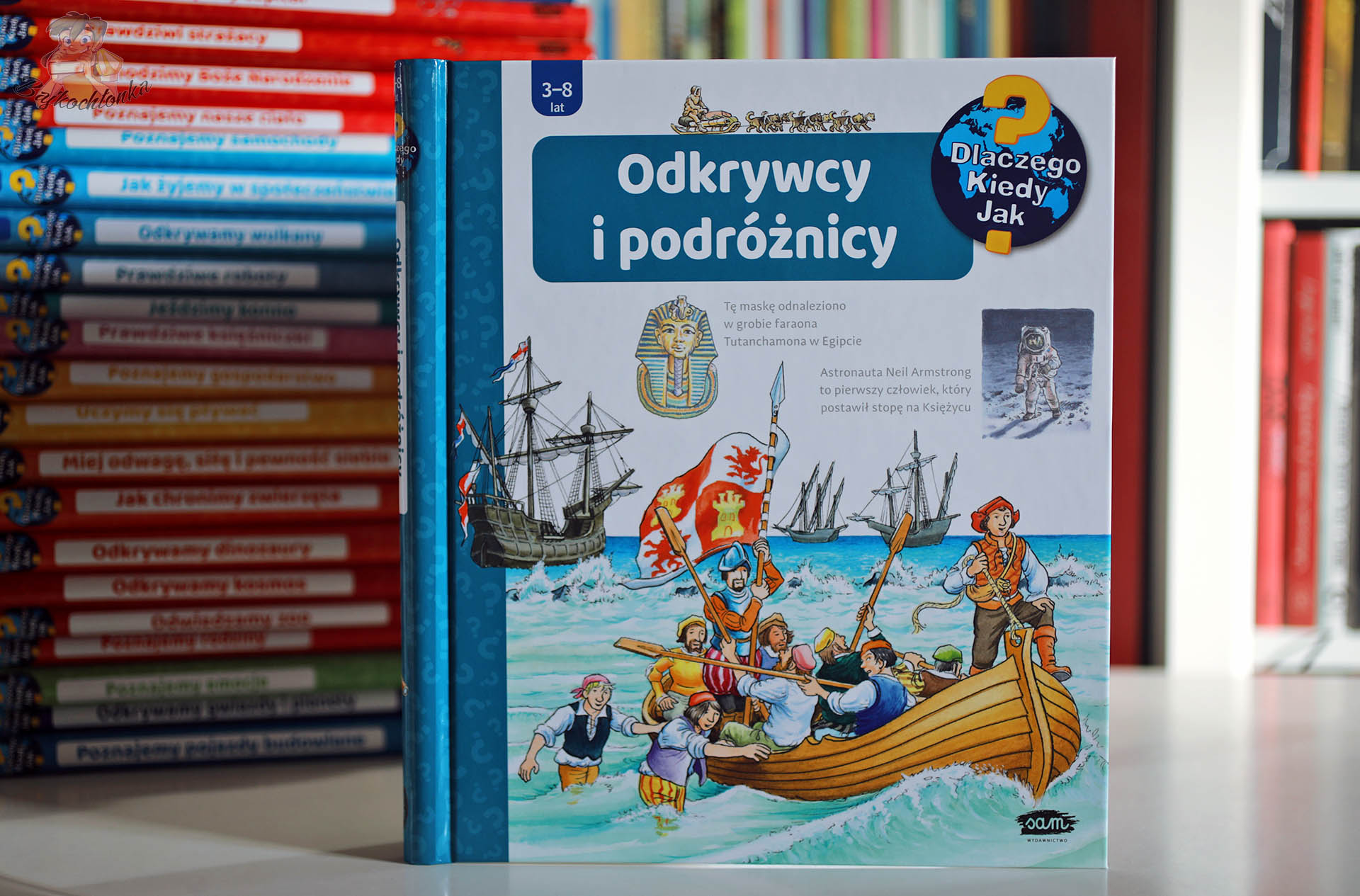 Okładka książki popularnonaukowej dla dzieci Odkrywcy i podróżnicy z serii Dlaczego? Kiedy? Jak? – scena lądowania żeglarzy na brzegu, statki na wzburzonym morzu i ilustracje historyczne.