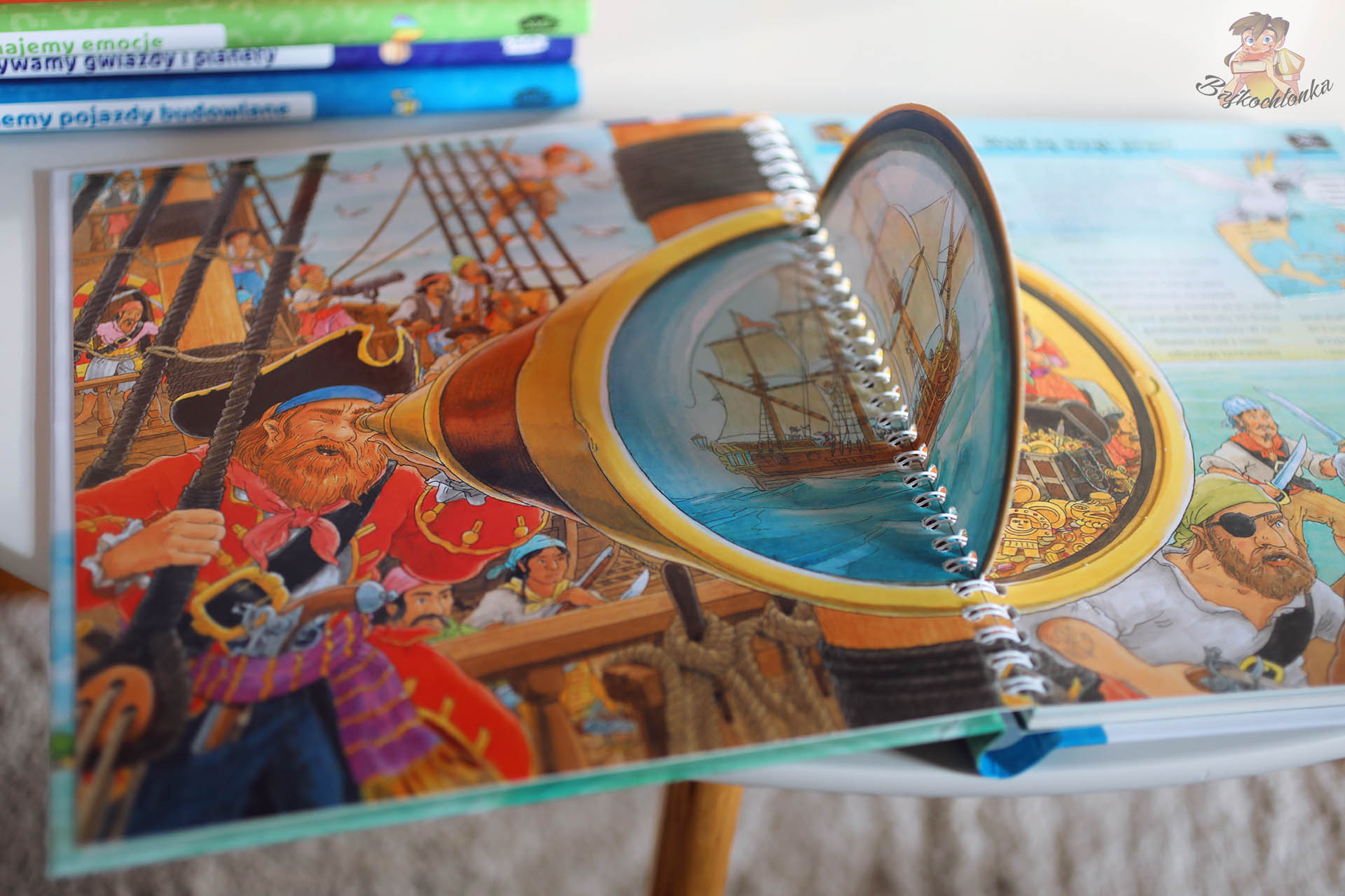 Rozkładówka z książki Prawdziwi piraci z wielkim trójwymiarowym okienkiem w kształcie pirackiej lunety, przez którą widać żaglowiec na morzu, otoczony scenami z załogą.