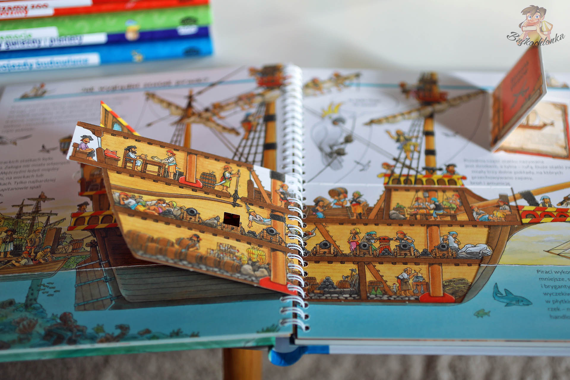 Otwarta książka Prawdziwi piraci z serii Dlaczego? Kiedy? Jak? pokazująca szczegółowy przekrój pirackiego statku z załogą, armatami i ładownią oraz dużymi, odchylonymi okienkami.