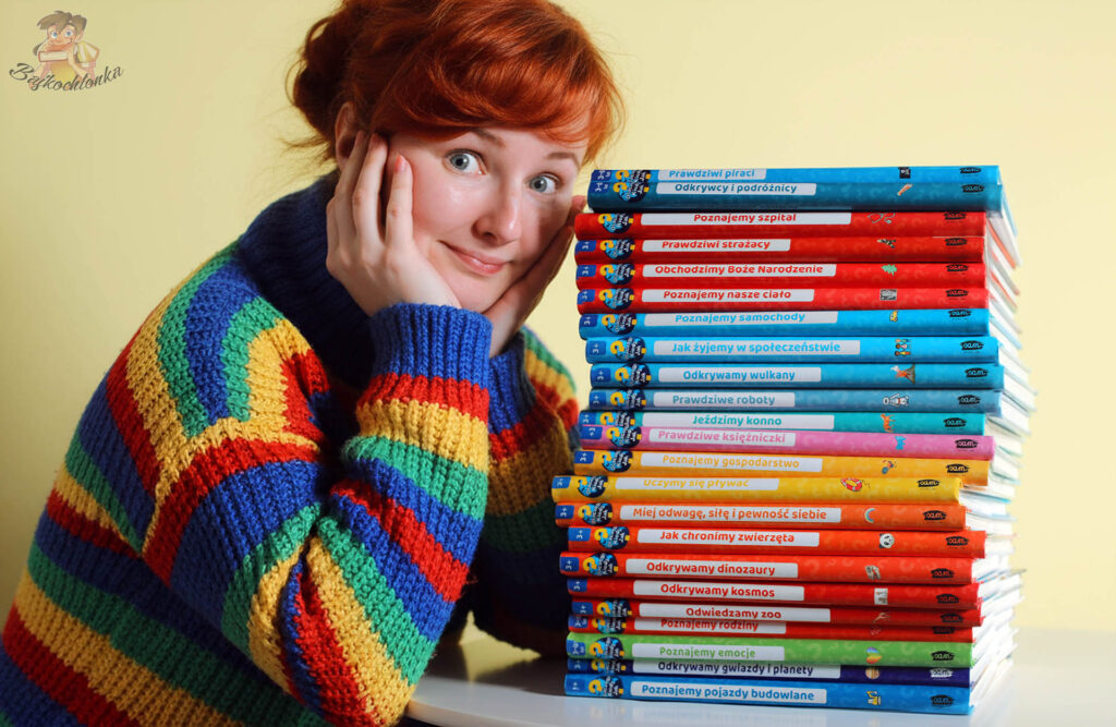 Uśmiechnięta osoba w kolorowym, tęczowym swetrze opiera się o wysoki stos książek popularnonaukowych dla dzieci z serii Dlaczego? Kiedy? Jak? ustawionych na żółtym tle.
