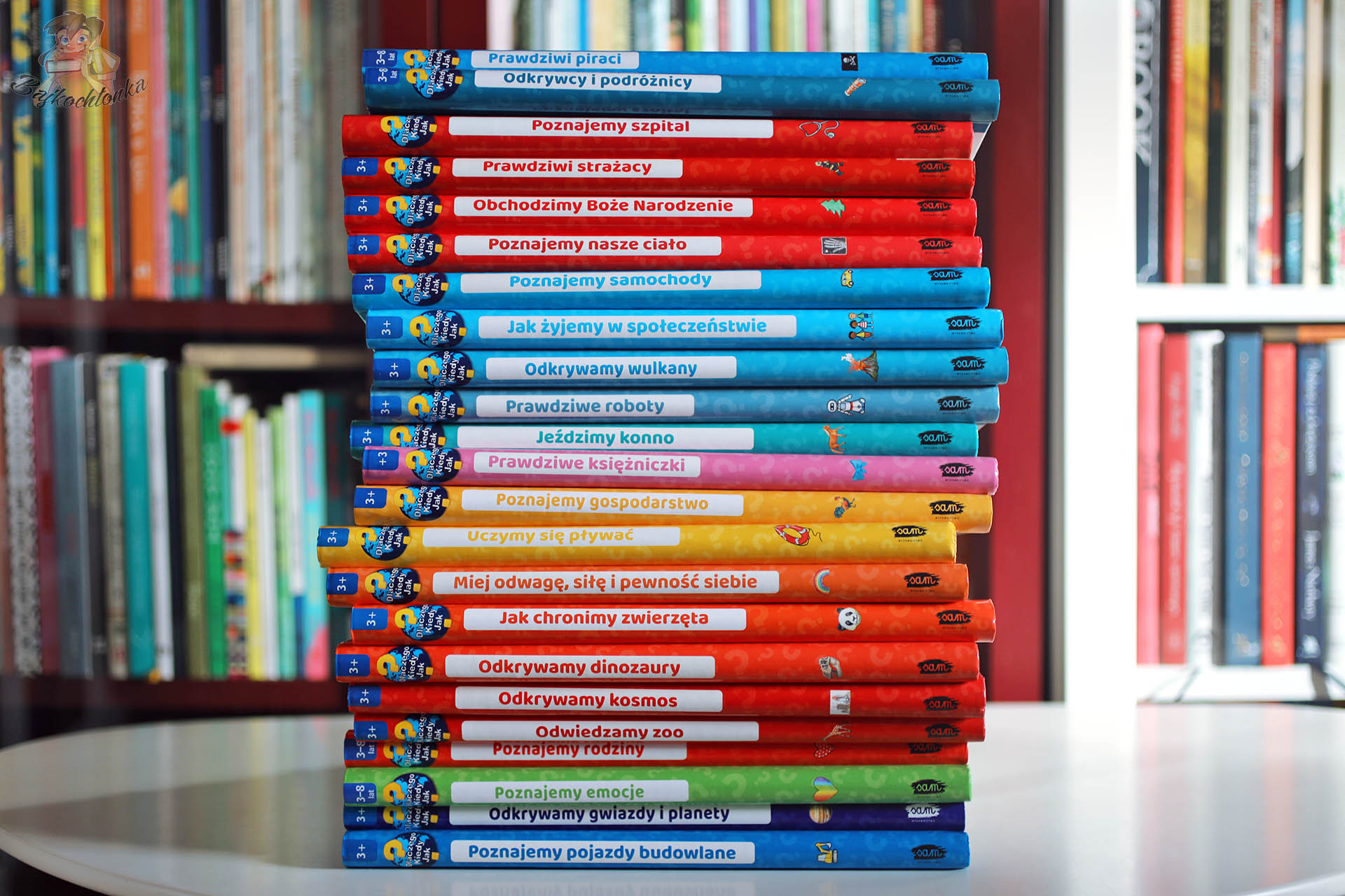 Wysoki stos kolorowych książek popularnonaukowych dla dzieci z serii Dlaczego? Kiedy? Jak? ustawiony na stole na tle regału z innymi tytułami w domowej biblioteczce.