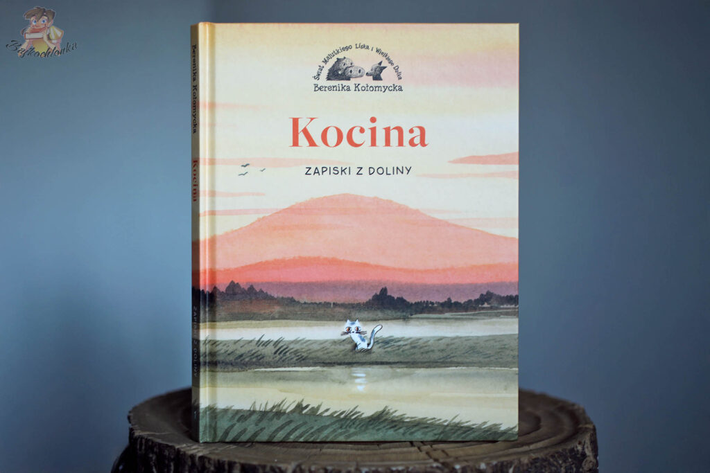 okładka komiksu Kocina. Zapiski z Doliny Bereniki Kołomyckiej z kotką nad wodą i pastelowym krajobrazem gór o zachodzie słońca