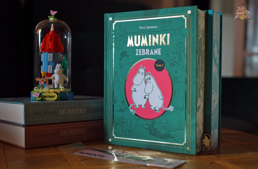 Stojący przodem tom 1 nowego, zielonego wydania Muminków zebranych od Naszej Księgarni, za nim ułożone poziomo dwa starsze pastelowe tomy oraz szklana kopuła z klockowym domkiem Muminków i figurką Muminka.