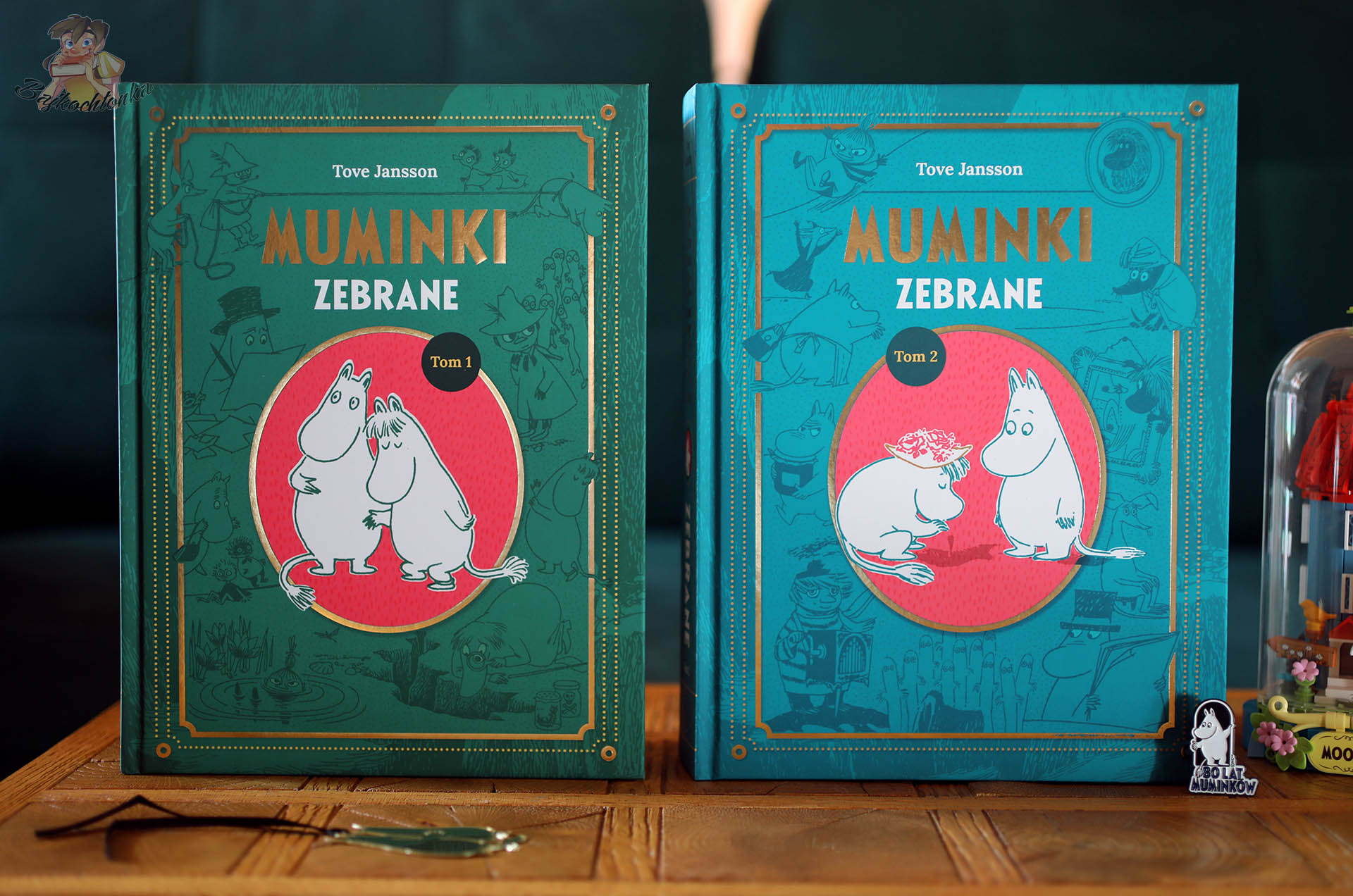 Dwa stojące obok siebie tomy 1 i 2 Muminków zebranych od Naszej Księgarni, w zielono–turkusowych okładkach ze złotymi zdobieniami i ilustracjami Muminków, na drewnianym stoliku z miniaturową budowlą z Muminkami po prawej stronie.