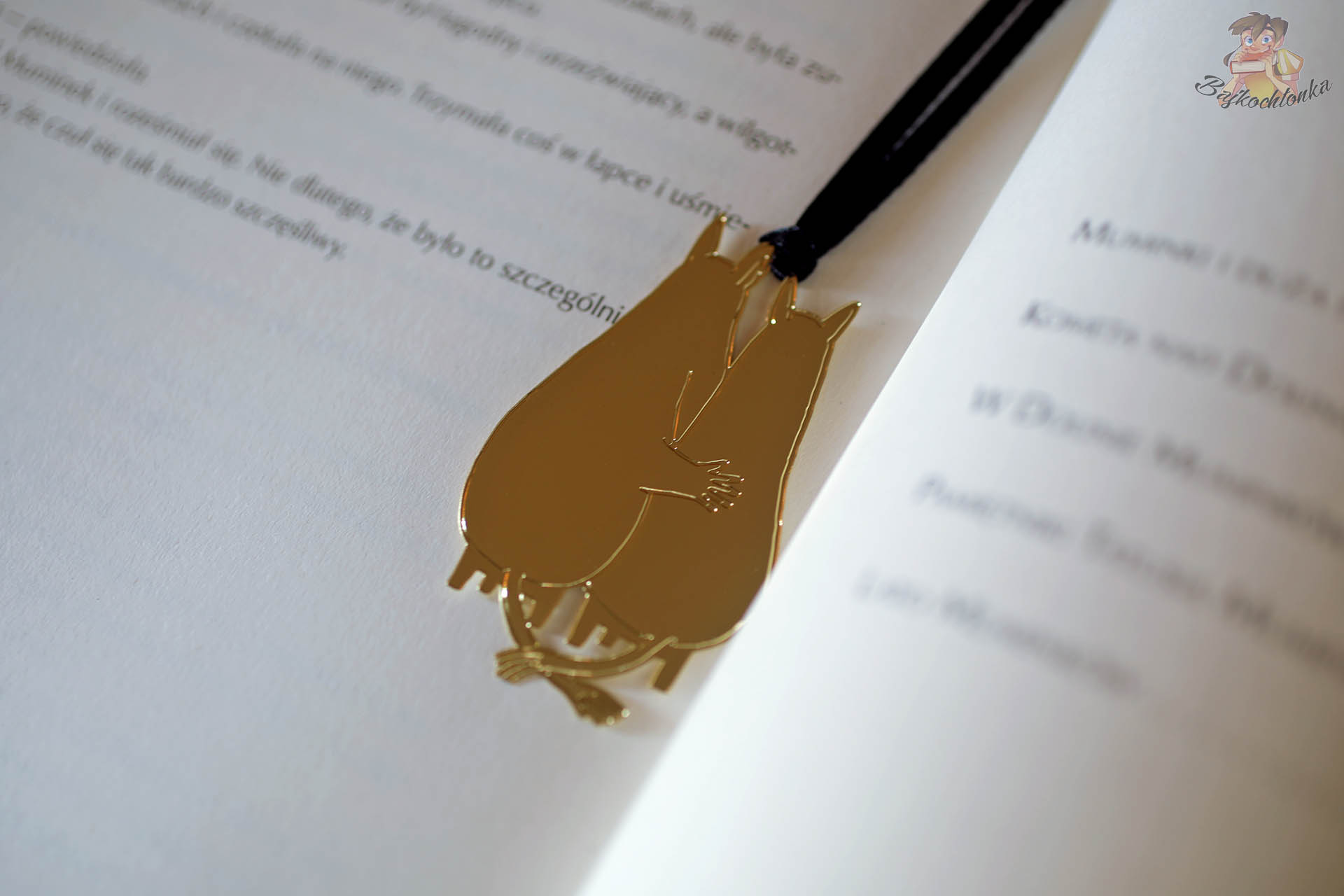 Metalowa, złota zakładka z obejmującymi się Muminkami leżąca między stronami nowego wydania Muminków zebranych od Naszej Księgarni.
