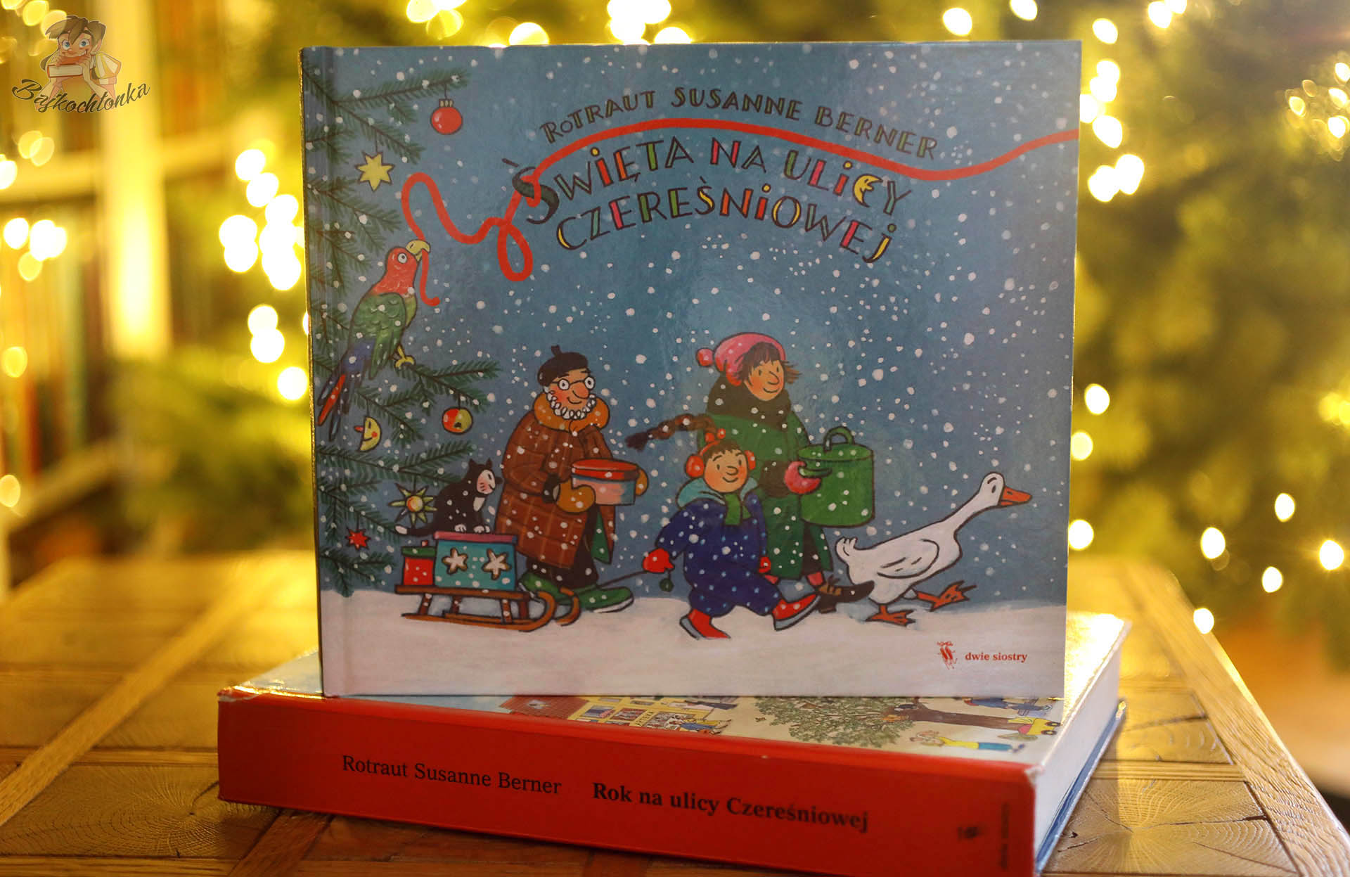 Okładka książki Święta na ulicy Czereśniowej ustawiona na tomie Rok na ulicy Czereśniowej na tle świątecznych światełek