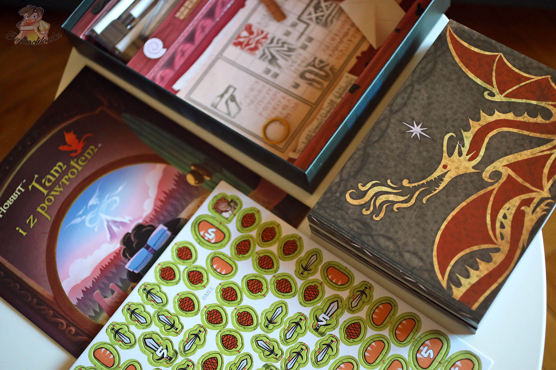 Żetony chleba i mieczy oraz stos książkowych plansz z wizerunkiem smoka ze Śródziemia z gry „Hobbit: Tam i z powrotem”