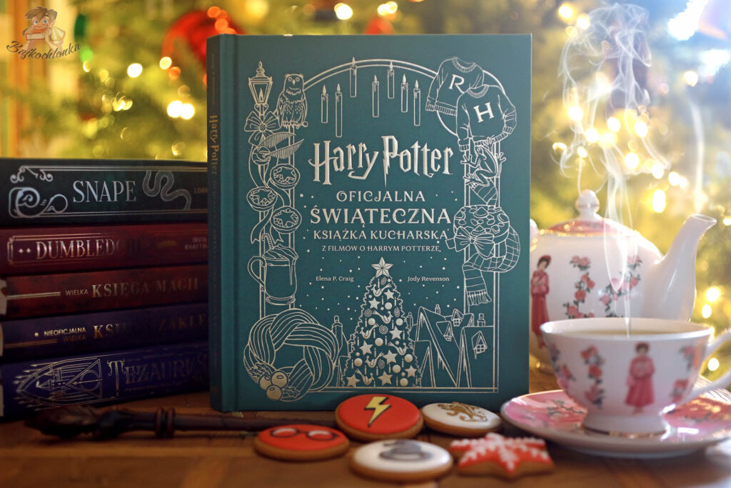 Książka Harry Potter. Oficjalna świąteczna książka kucharska stojąca przed choinką, otoczona potterowskimi tomami, filiżanką z herbatą i świątecznymi ciastkami z motywami z serii.