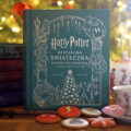 Książka Harry Potter. Oficjalna świąteczna książka kucharska stojąca przed choinką, otoczona potterowskimi tomami, filiżanką z herbatą i świątecznymi ciastkami z motywami z serii.