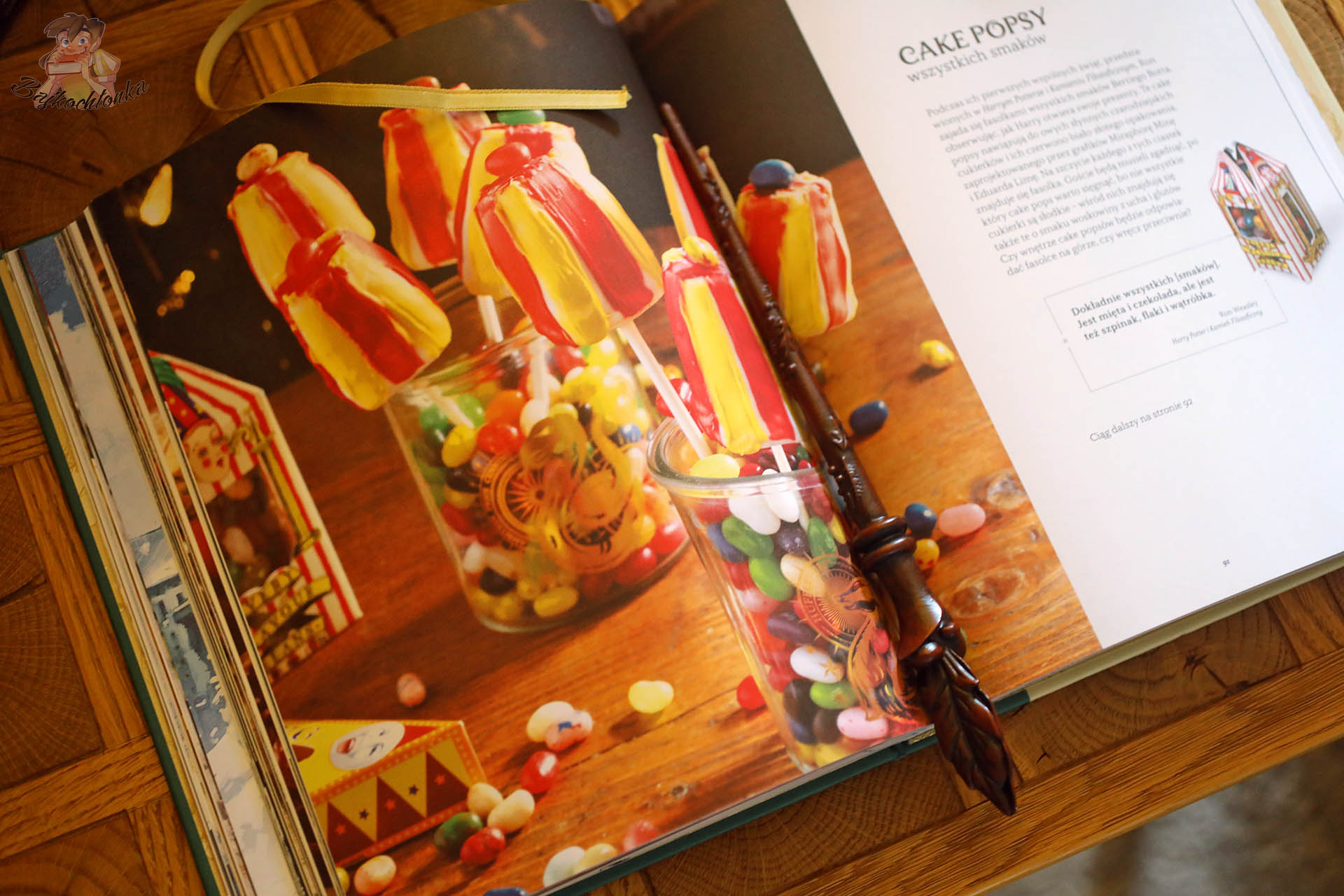 Rozłożona Harry Potter. Oficjalna świąteczna książka kucharska z kolorowymi cake popsami inspirowanymi słodyczami z Hogwartu, słoikami z fasolkami i różdżką leżącą na stronach.