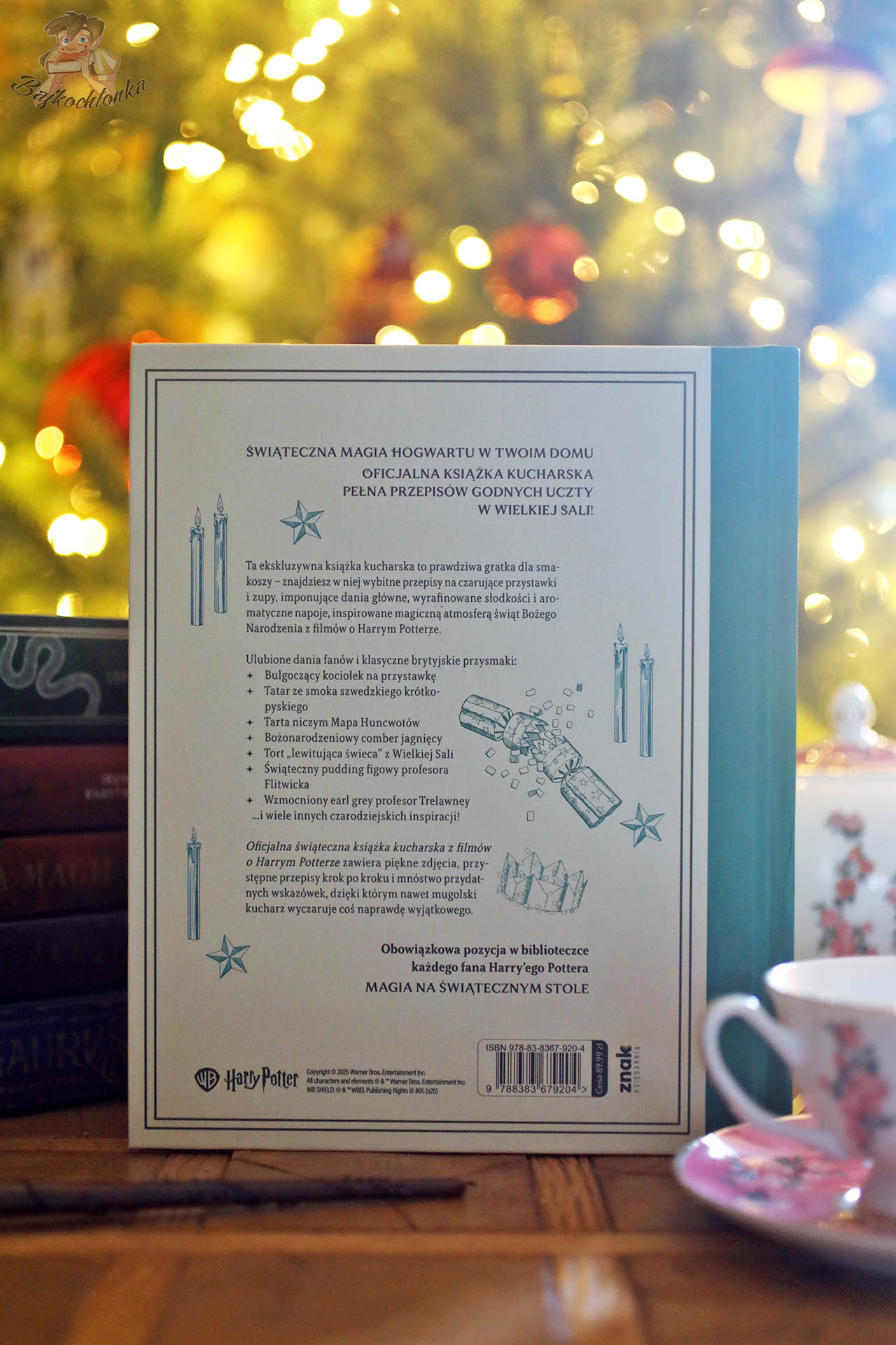 Tył okładki Harry Potter. Oficjalna świąteczna książka kucharska z opisem „Świąteczna magia Hogwartu w twoim domu”, sfotografowany na tle rozświetlonej choinki i świątecznych ozdób.