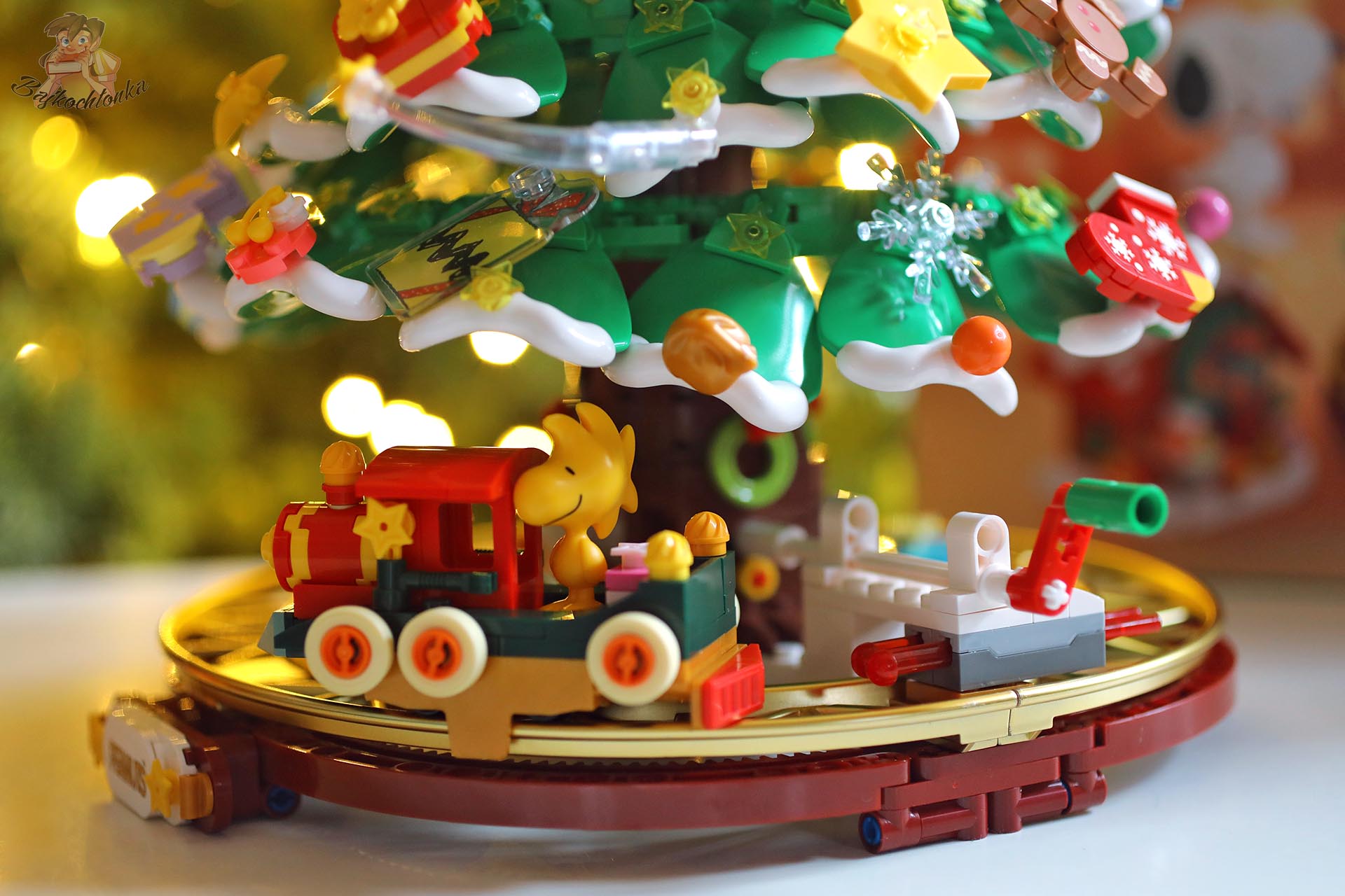 Pociąg Woodstocka jeżdżący po złotych torach w zestawie Pantasy Peanuts Christmas Tree 86909, z ozdobami u podstawy choinki.