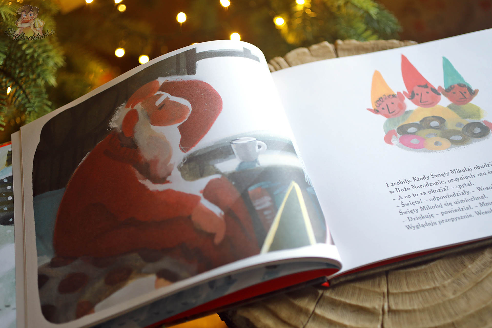 Zbliżenie na rozłożoną książkę Pierwsza Gwiazdka Świętego Mikołaja z ilustracją siedzącego przy stole Świętego Mikołaja oraz trzema uśmiechniętymi elfami z talerzem pączków, w tle choinka.
