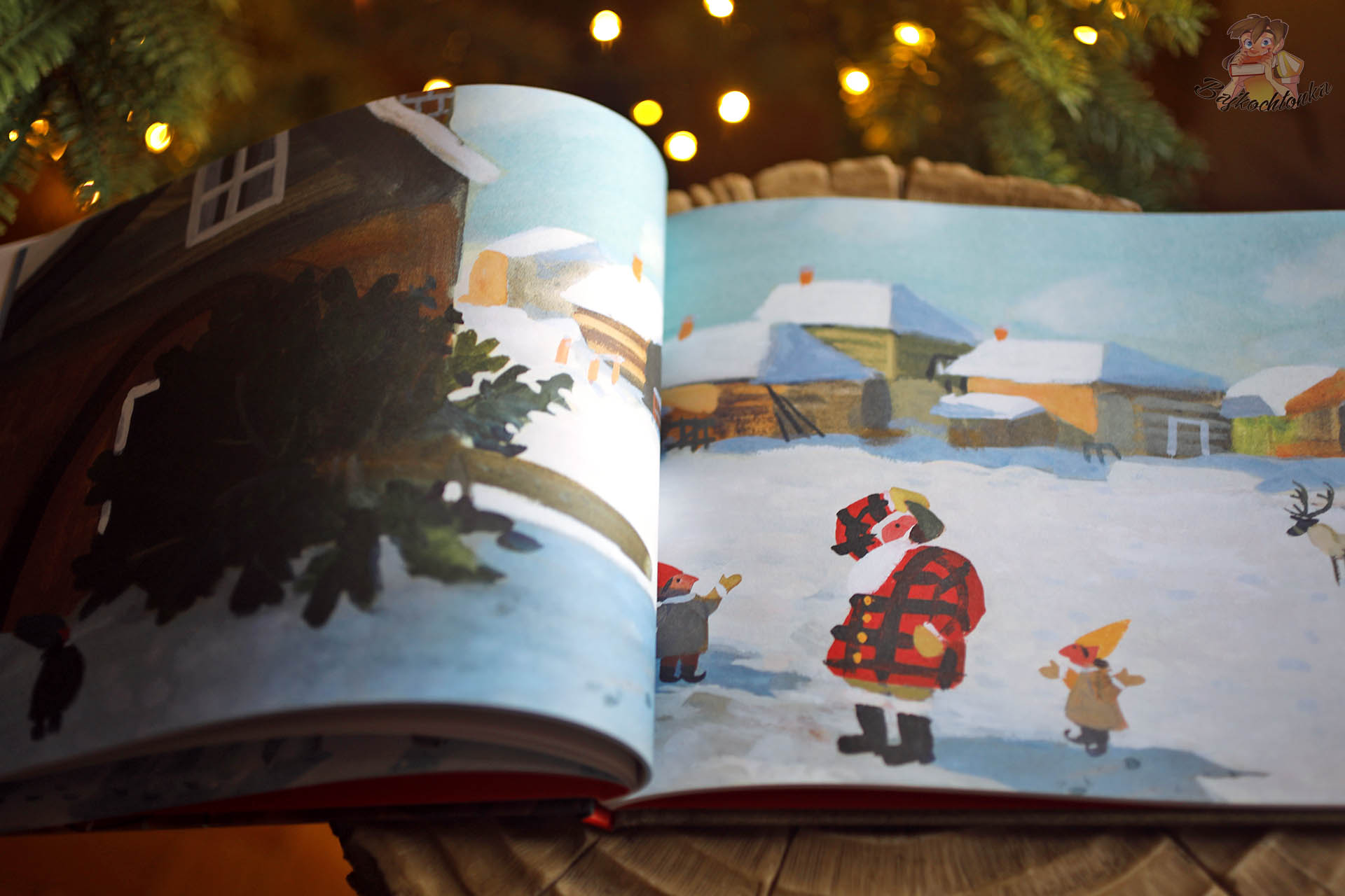 Strony książki Pierwsza Gwiazdka Świętego Mikołaja z ilustracją Świętego Mikołaja i elfów stojących na ośnieżonym podwórzu między domkami, w tle choinka i światełka.