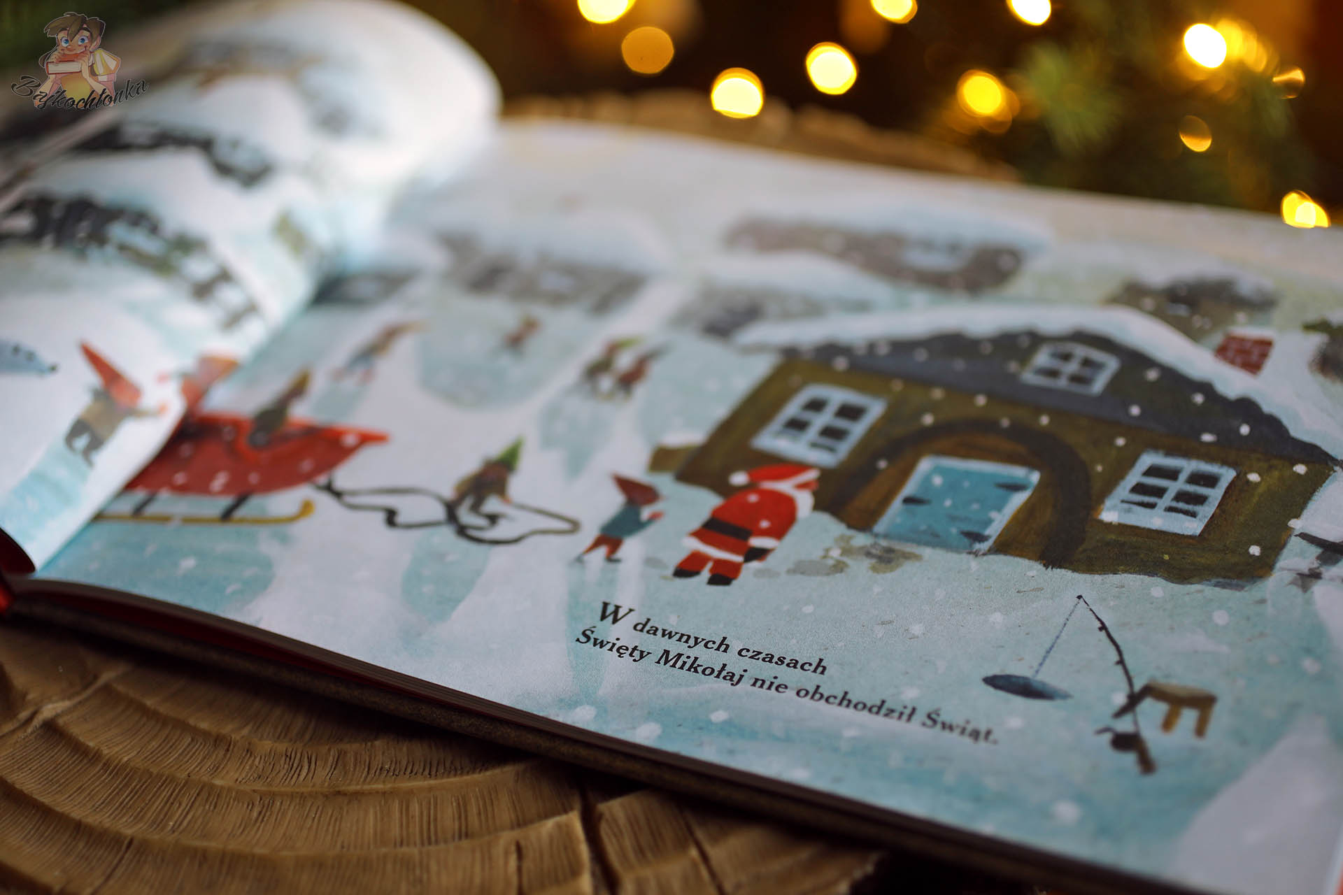 Otwarta książka Pierwsza Gwiazdka Świętego Mikołaja z zimową sceną miasteczka, na pierwszym planie Mikołaj i elf idący w stronę domu oraz widoczny fragment tekstu „W dawnych czasach Święty Mikołaj nie obchodził Świąt”.