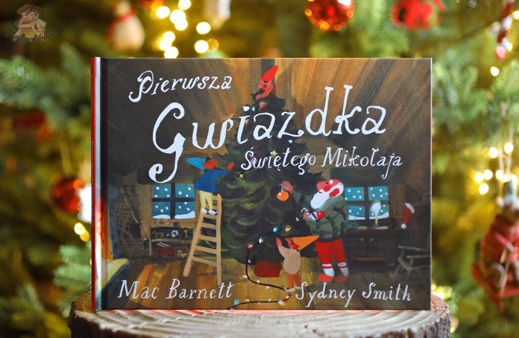 Książka Pierwsza Gwiazdka Świętego Mikołaja stojąca na drewnianym pieńku przed choinką, na okładce elfy i Święty Mikołaj dekorujący świąteczne drzewko w ciepłym, przytulnym wnętrzu.