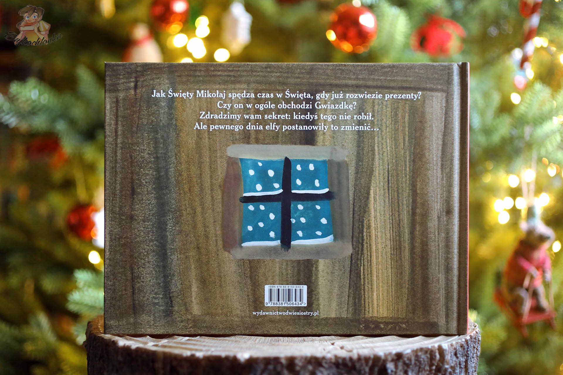 Tył okładki książki Pierwsza Gwiazdka Świętego Mikołaja z tekstem zapowiadającym historię i namalowanym oknem z płatkami śniegu, ustawiony na drewnianym pieńku przed świąteczną choinką.