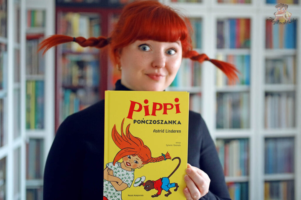 Bajkochłonka z rudymi włosami upiętymi jak u Pippi trzyma przed sobą książkę Pippi Pończoszanka w żółtej okładce, w tle widać regały pełne książek.