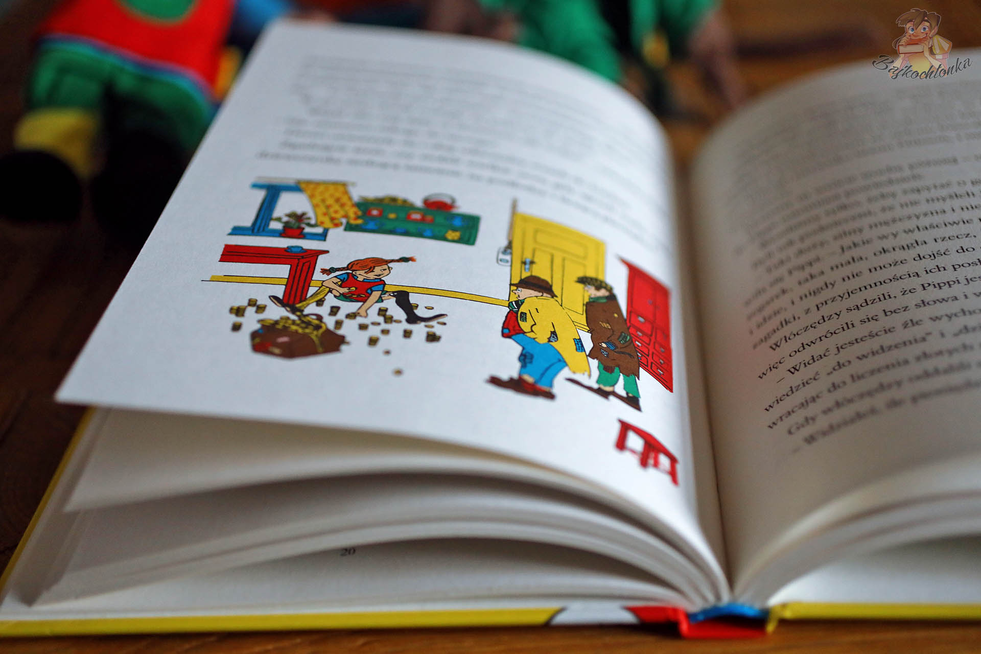 Otwarte strony Pippi Pończoszanki z ilustracją, na której Pippi rozsypuje złote monety przed zaskoczonymi złodziejami stojącymi w drzwiach Willi Śmiesznotki.