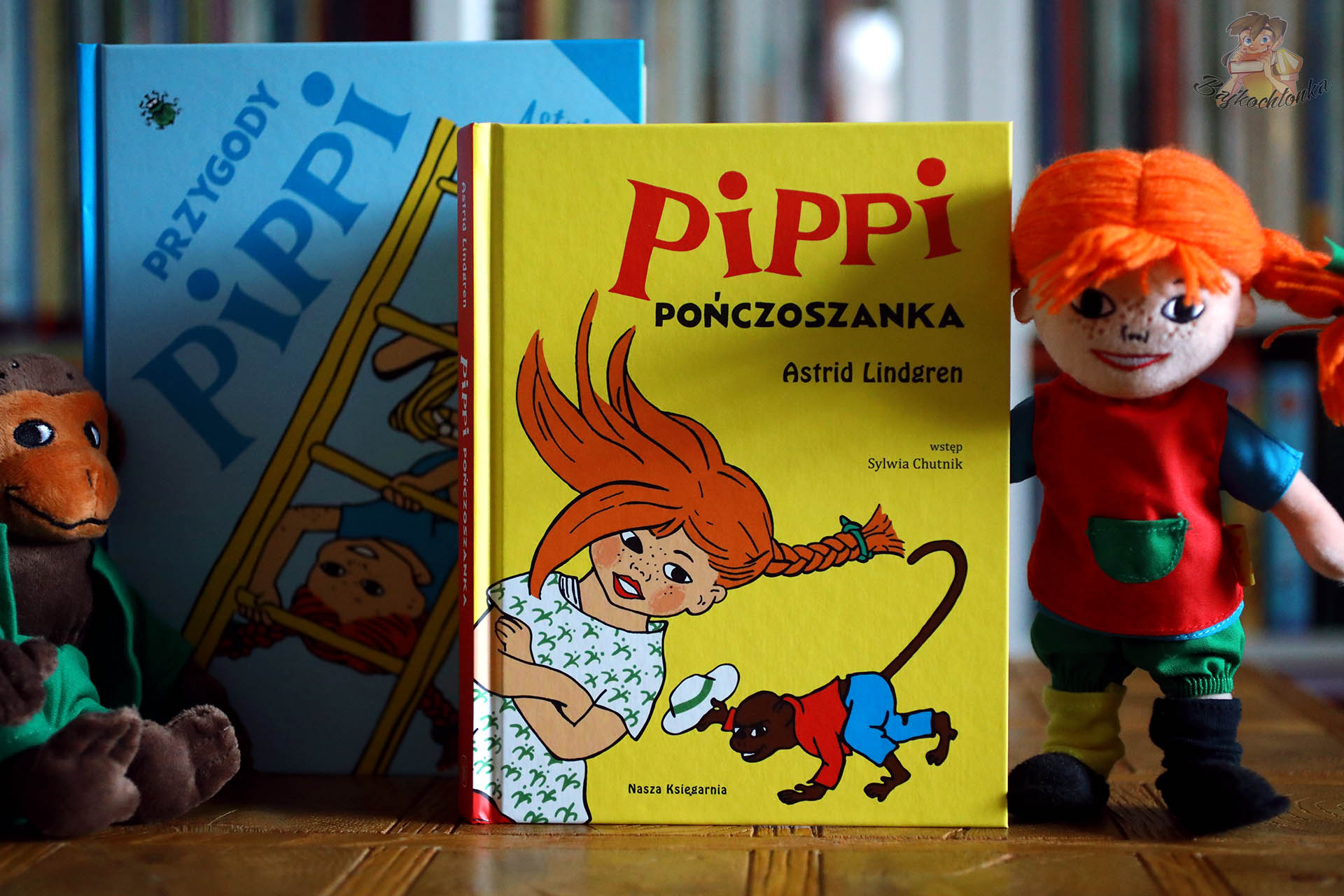 Okładka książki Pippi Pończoszanka w żółtej oprawie ustawiona przed niebieskim tomem Przygody Pippi, po bokach stoją pluszowe postaci Pippi i małpki.