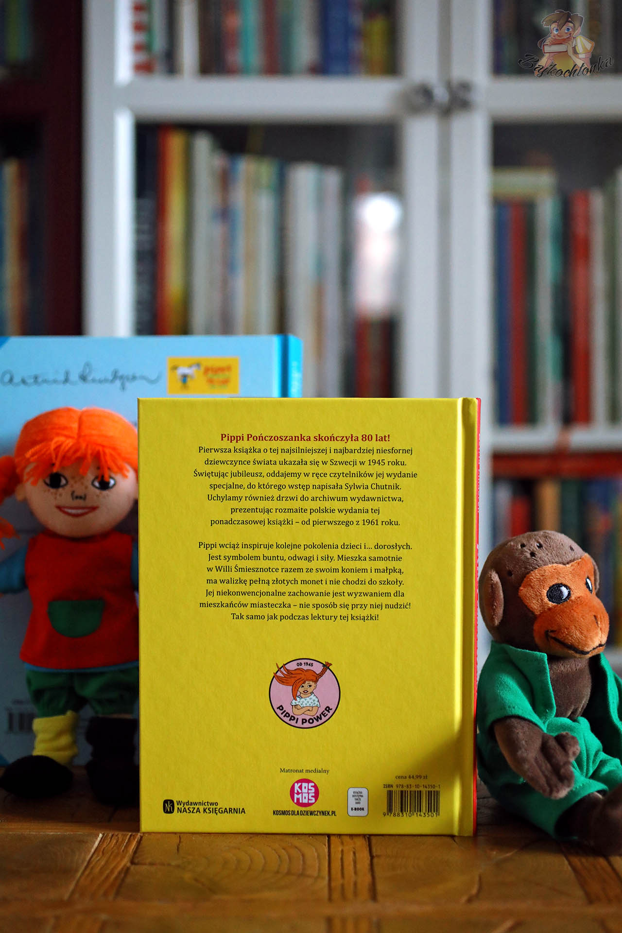 Tył żółtej okładki książki Pippi Pończoszanka ustawionej na tle regału z książkami, obok stoją pluszowe zabawki Pippi i małpki Pana Nilsona.