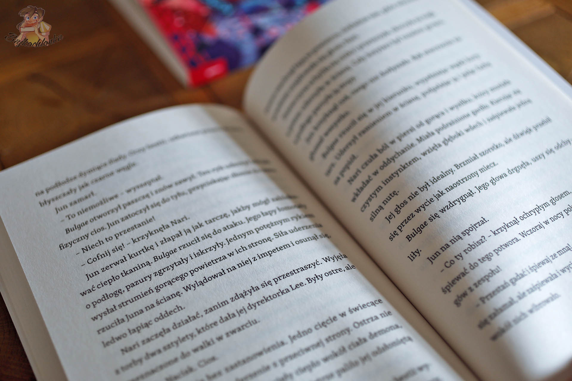 Fragment powieści „Scena cieni” z serii K-Pop Academy autorstwa Mina Finch – otwarta książka z widocznym dialogiem i sceną walki z demonem