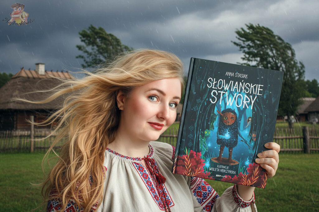 Książka Słowiańskie stwory Anny Stasiak trzymana w dłoni na tle słowiańskiej wioski