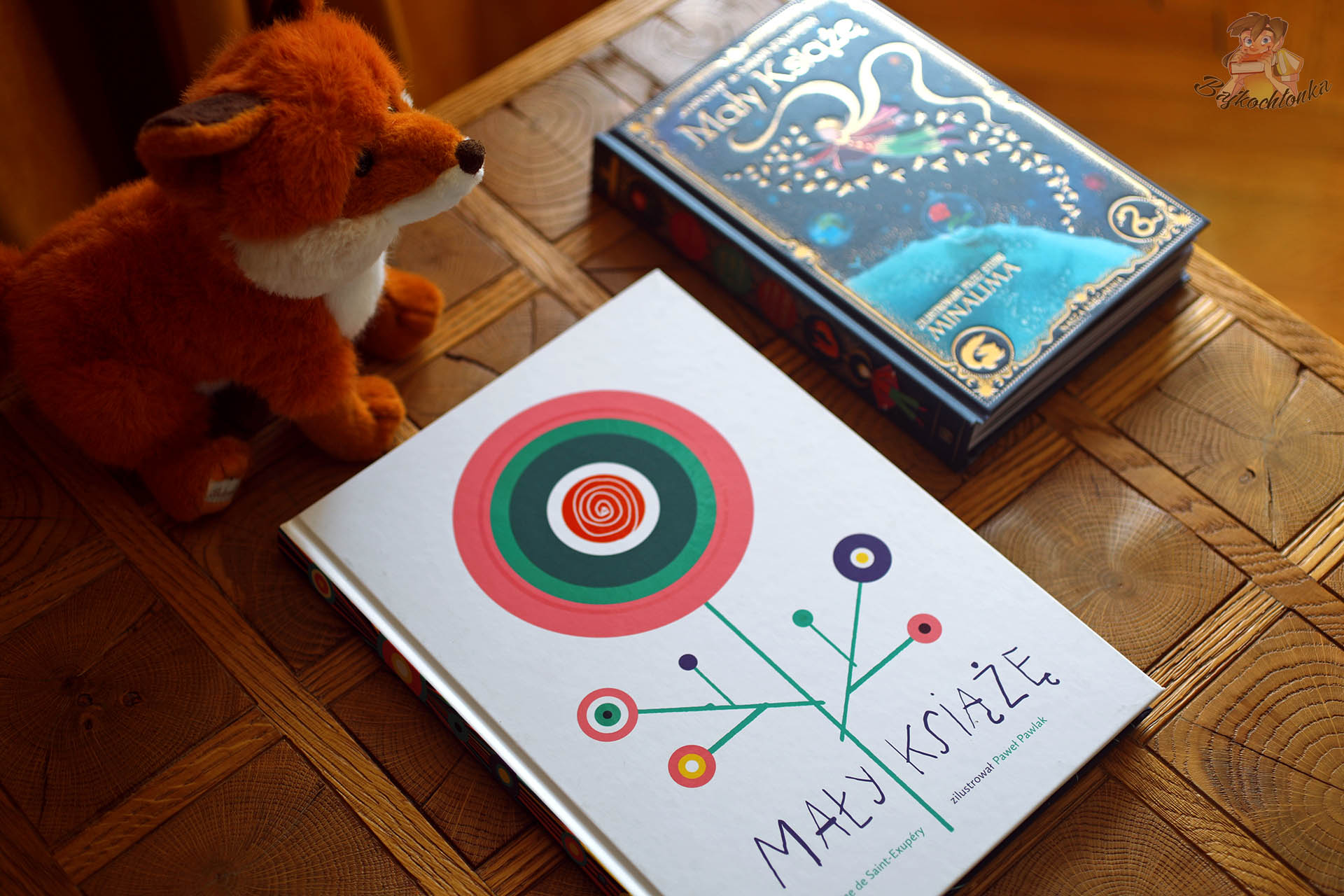dwa wydania Małego Księcia na drewnianym stole – edycja MinaLima i wydanie z ilustracjami Pawła Pawlaka z pluszowym liskiem
