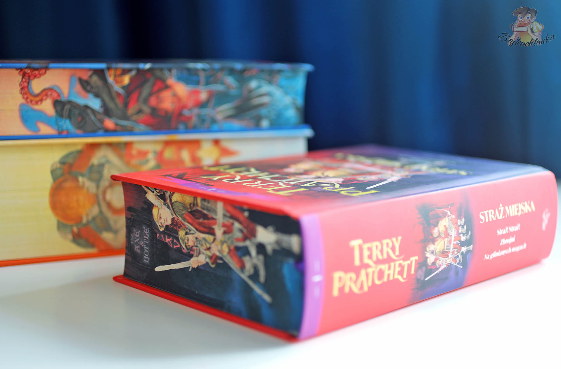 Grzbiet książki Straż miejska. Tom 1 (barwione brzegi) na tle tomów Rincewind. Tom 1 i Rincewind. Tom 2