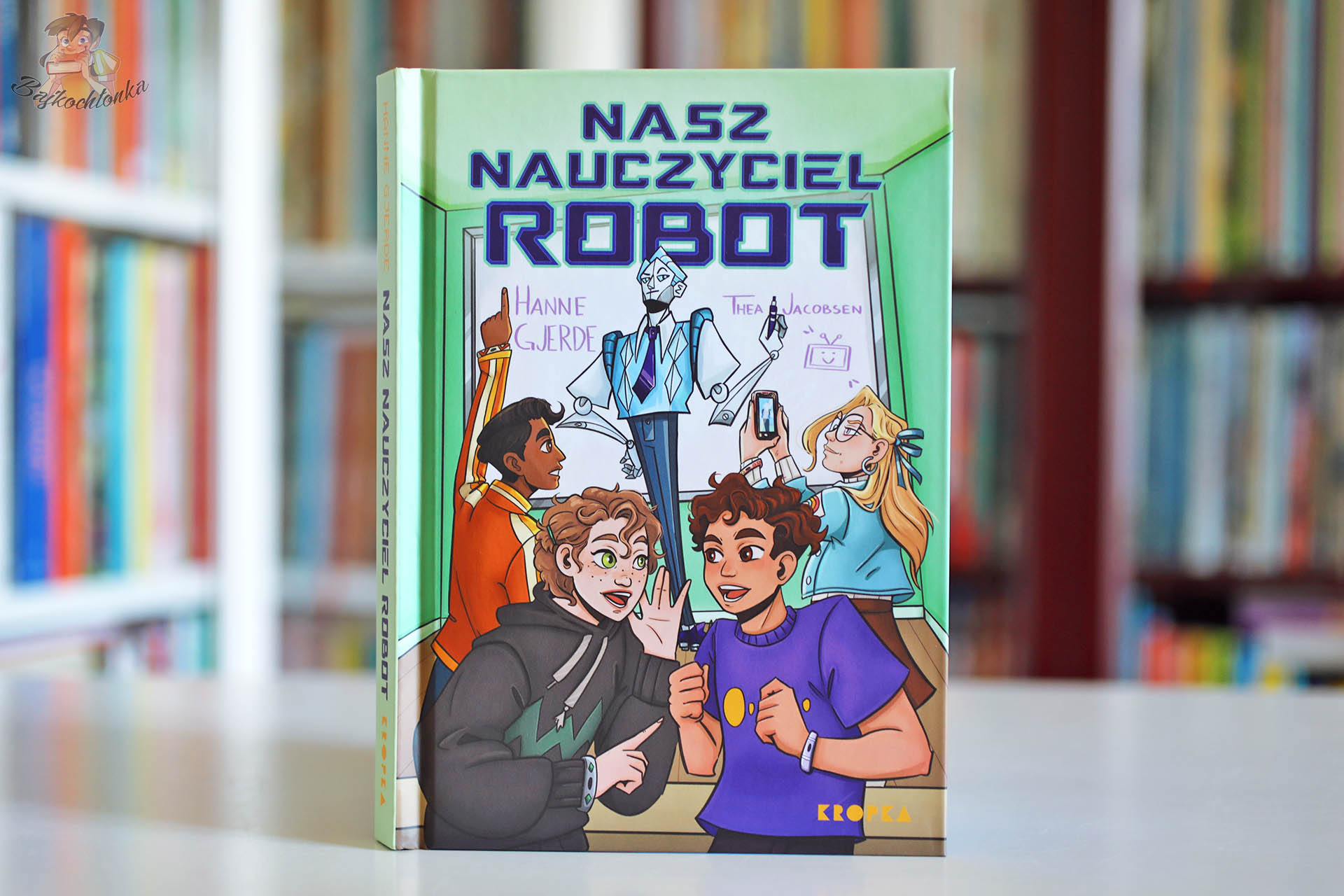 Okładka książki Nasz nauczyciel robot Hanne Gjerde i Thei Jacobsen wydanej przez Kropkę na tle biblioteczki