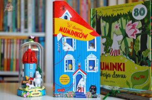 Okładka książki W domu Muminków z otwieranymi okienkami, stojąca obok muminkowej figurki i książki pop-up