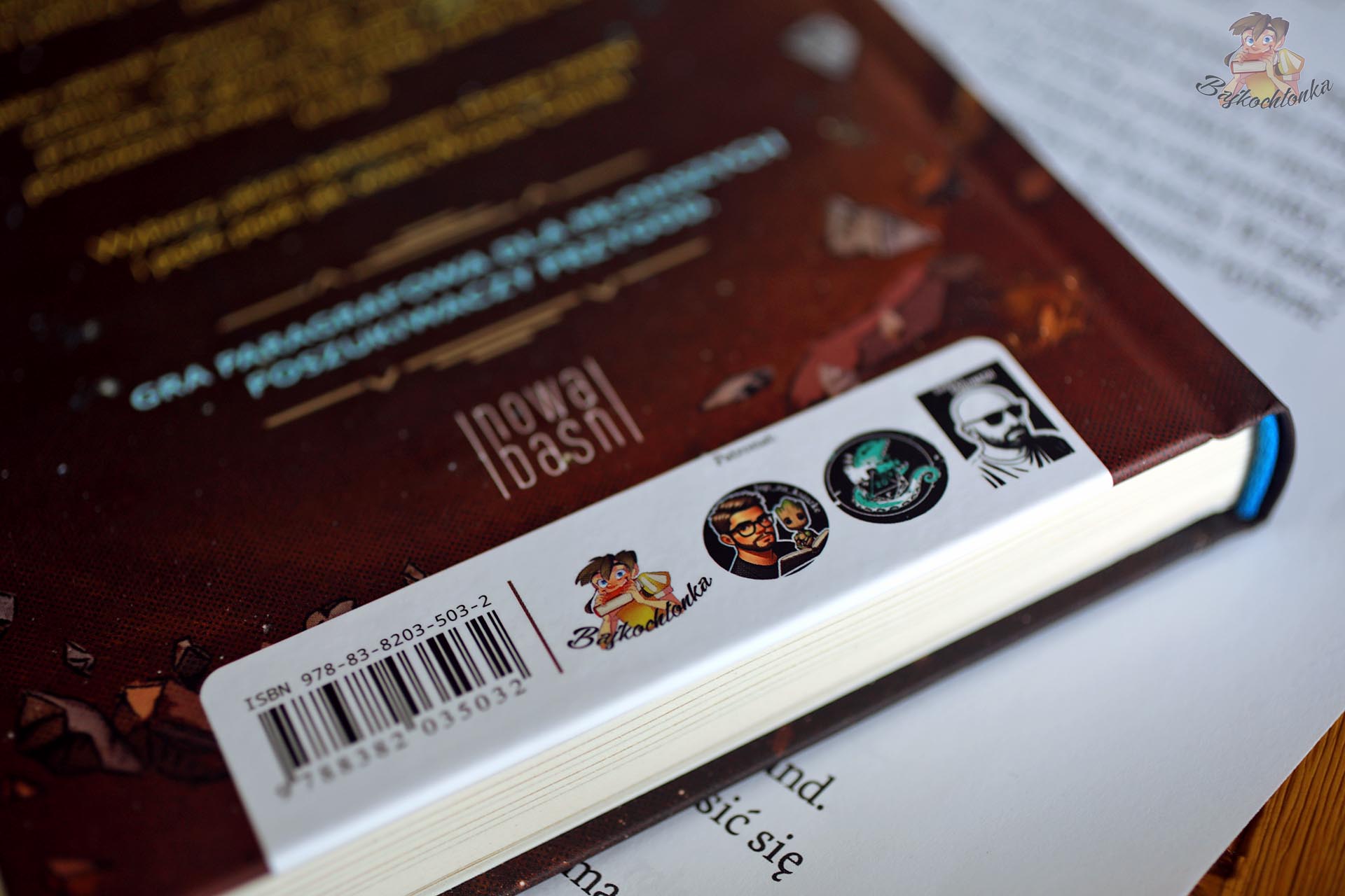 Zbliżenie na egzemplarz książki Wrona Cienia wydanej przez Nową Baśń, z widocznym grzbietem, kodem ISBN i oznaczeniami patronów.