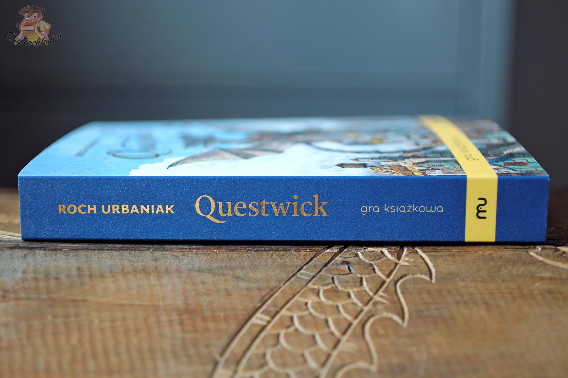 Grzbiet gry książkowej „Questwick. Stolica Przygody” Rocha Urbaniaka wydanej przez Muduko