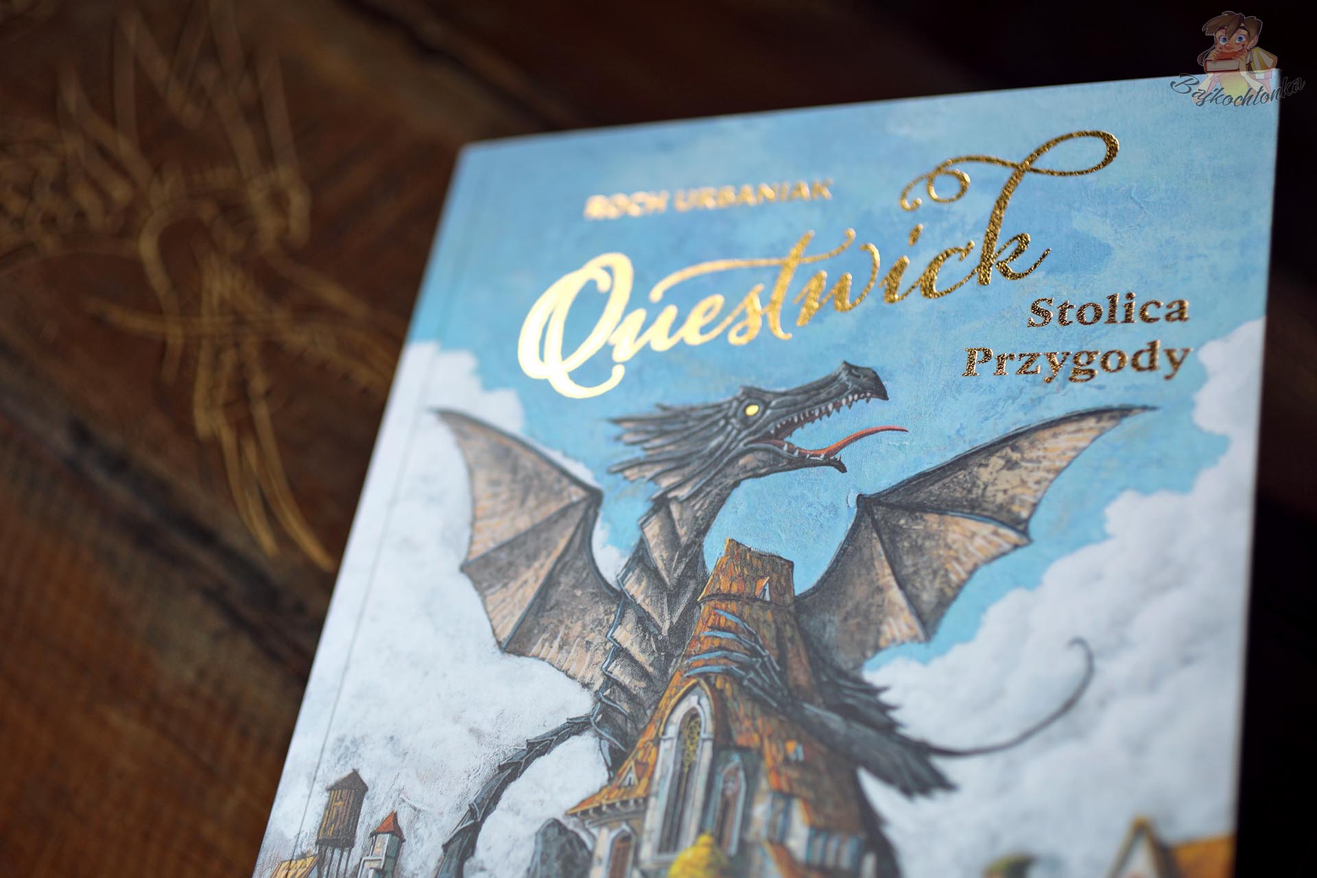 Złote uszlachetnienia, połyskujący tytuł i oko smoka na okładce książki „Questwick. Stolica Przygody”