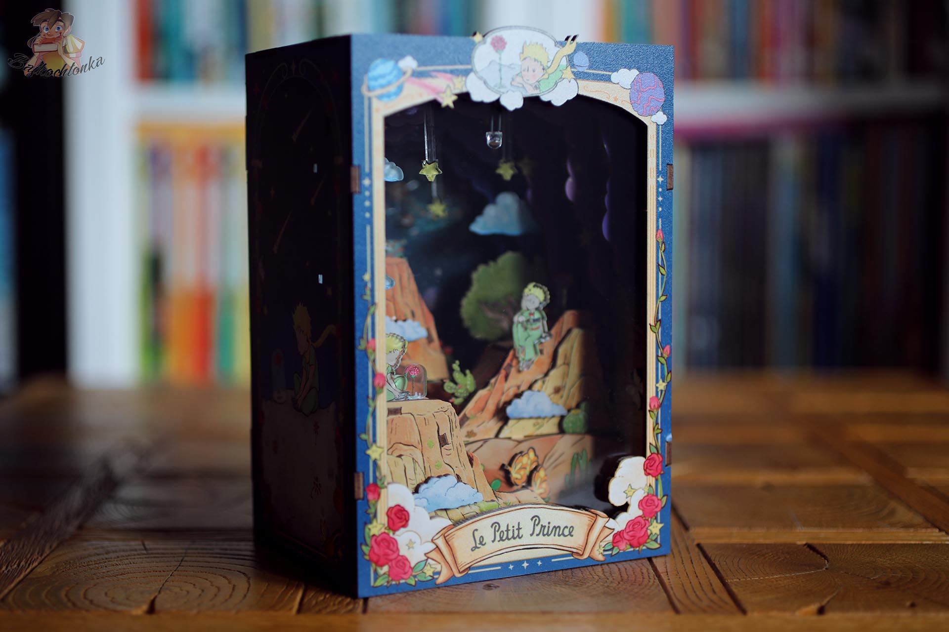 Gotowy book nook Mały Książę i Róża ustawiony pod kątem na tle regału z książkami
