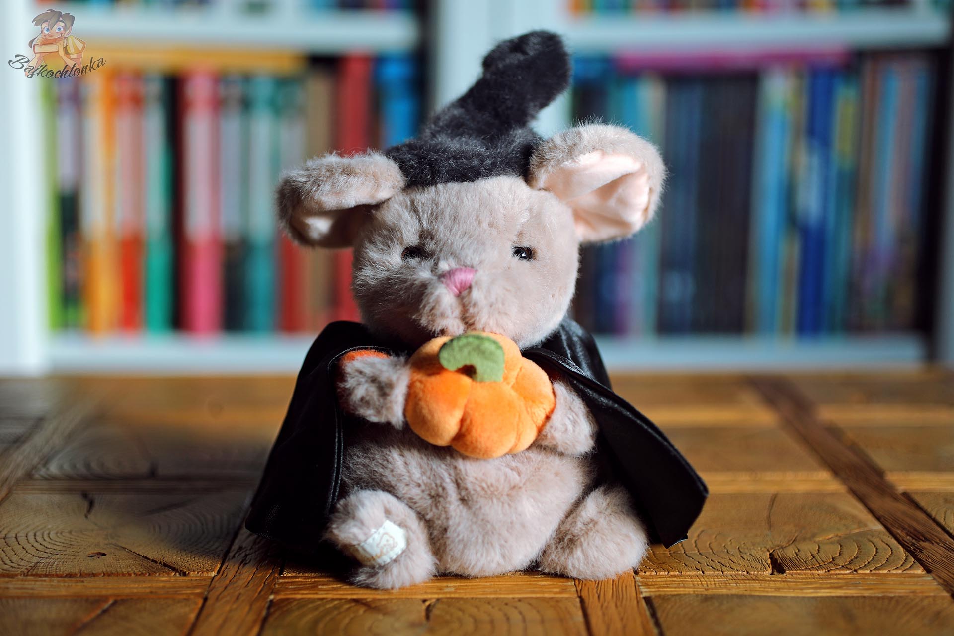 Zbliżenie na pluszową myszkę Halloween Mousy w czarnym kapeluszu i pelerynie, trzymającą pomarańczową dynię.