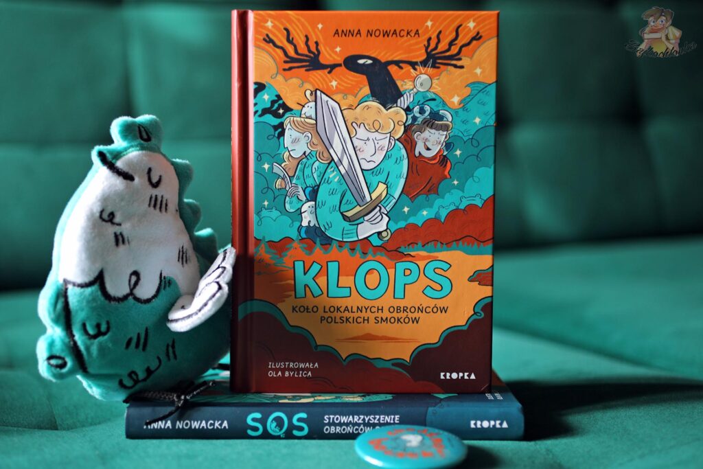 KLOPS. Koło Lokalnych Obrońców Polskich Smoków Anny Nowackiej – okładka książki z ilustracją Oli Bylicy, obok pluszowy smok i pierwszy tom SOS.