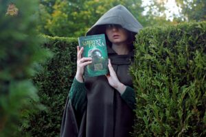 Postać w ciemnym kapturze trzyma książkę Labirynt Fauna Cornelii Funke i Guillermo del Toro wśród zielonych żywopłotów przypominających labirynt.