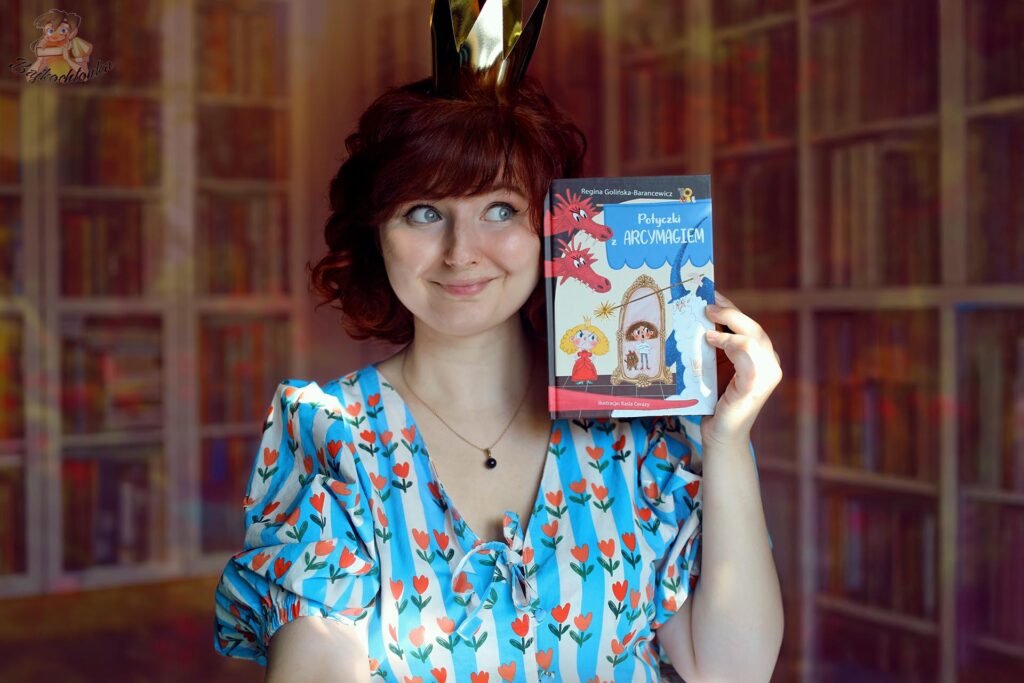 Kobieta w królewskiej koronie prezentuje książkę Potyczki z Arcymagiem Reginy Golińskiej-Barancewicz na tle domowej biblioteczki.